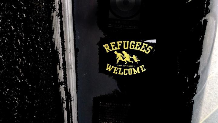 shiny black canister vandalized with a few stickers, one reading 'Refugees Welcome' with a yellow outline of two parents and child running away