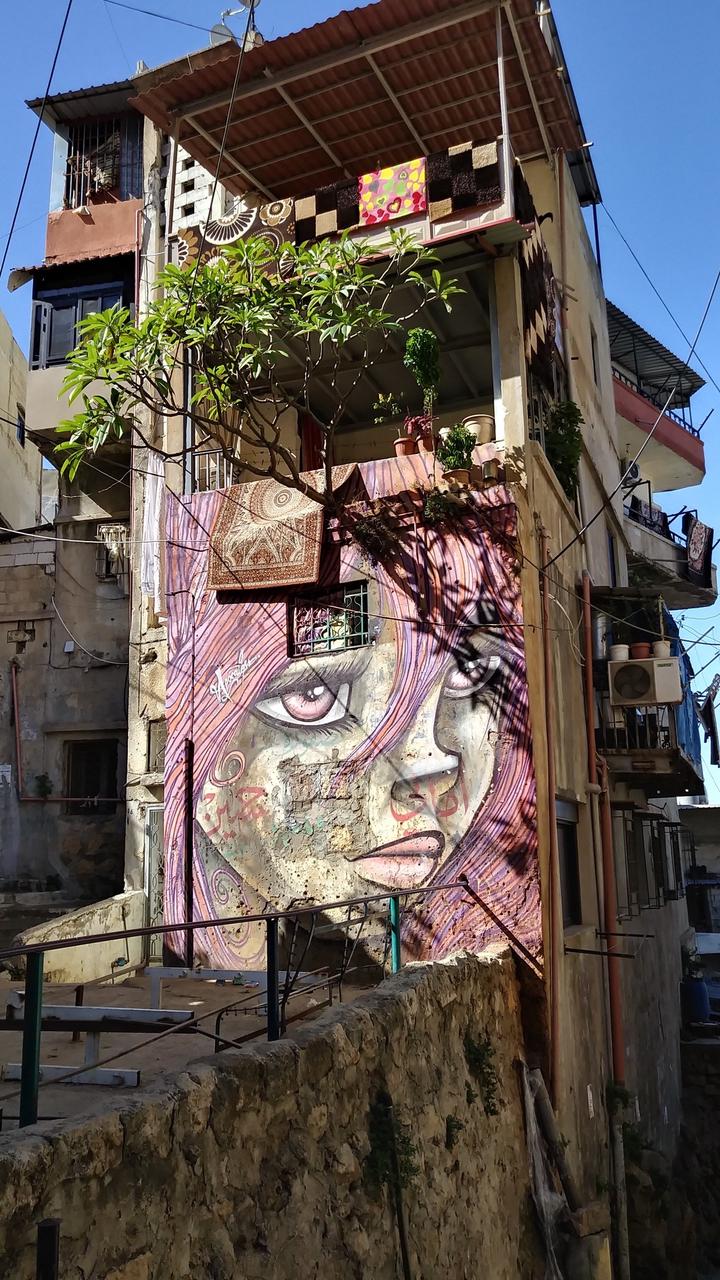 Lebanese apartment building on a clear day, the bottom wall is painted with graffiti art of a girl with pink-purple hair with lavender eyes staring absently
